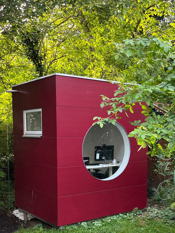 Kleines Gartenbuero in Kubusform mit Holzfassade in Rot, rundem Fenster, Lueftungsfenster mitten im gruenen Garten