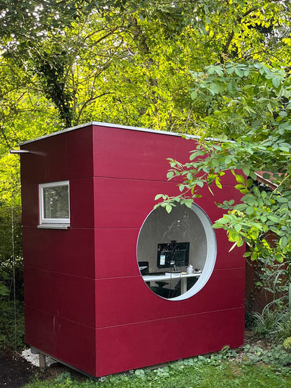 Kleines Gartenbuero in Kubusform mit Holzfassade in Rot, rundem Fenster, Lueftungsfenster mitten im gruenen Garten